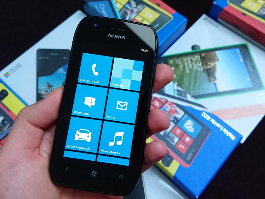 Ręka trzymająca czarną Nokię Lumię 710 z wieloma pudełkami z serii Nokia Lumia w tle
