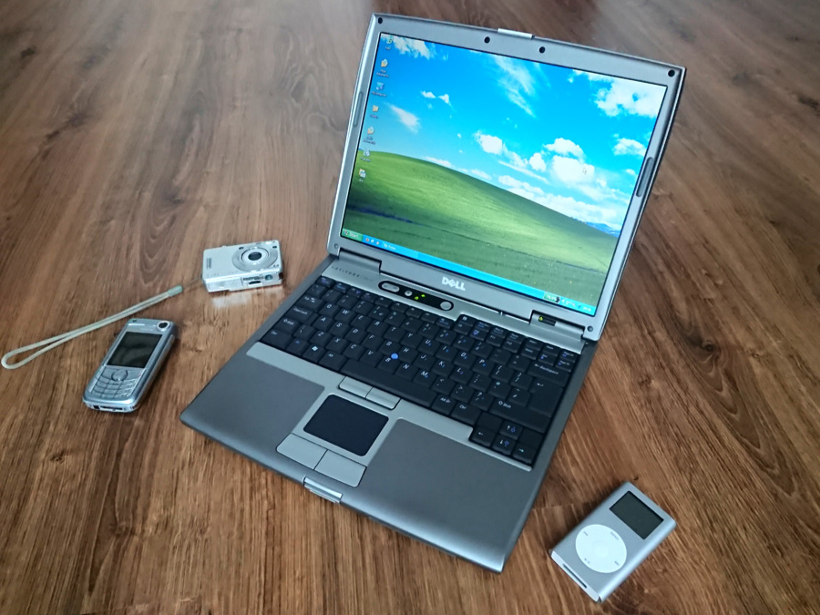 Four silver devices from around 2005 arranged on the floor. From left: a Nokia 6680 smartphone, Sony Cyber-shot DSC-W50 digital camera, Dell Latitude D610 laptop and Apple iPod mini music player